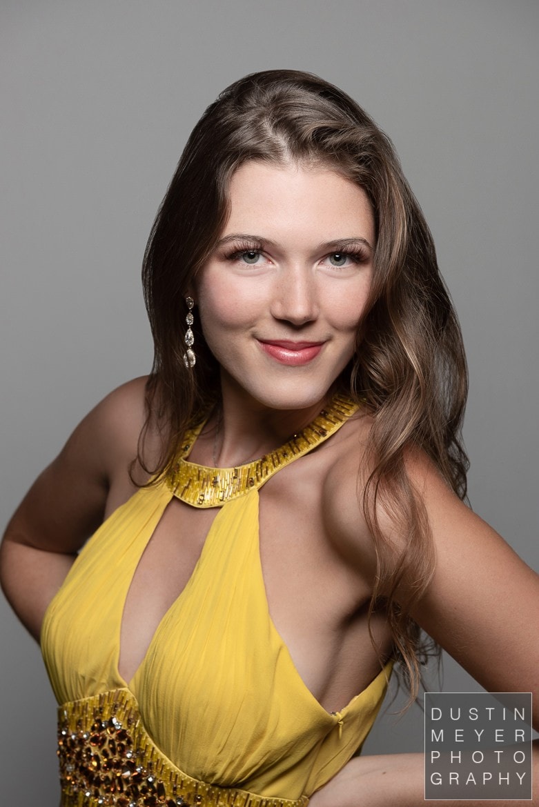 A brunette female model in a professional photography studio wearing a yellow dress in front of a white backdrop indoors