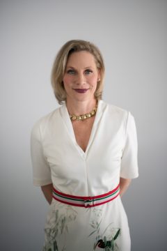 A professional company headshot of a blonde woman wearing a white dress in front of a white background with natural light.